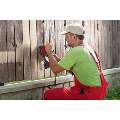 A man in red workwear is sanding a wood veneer with an electric polishing machine outdoors.