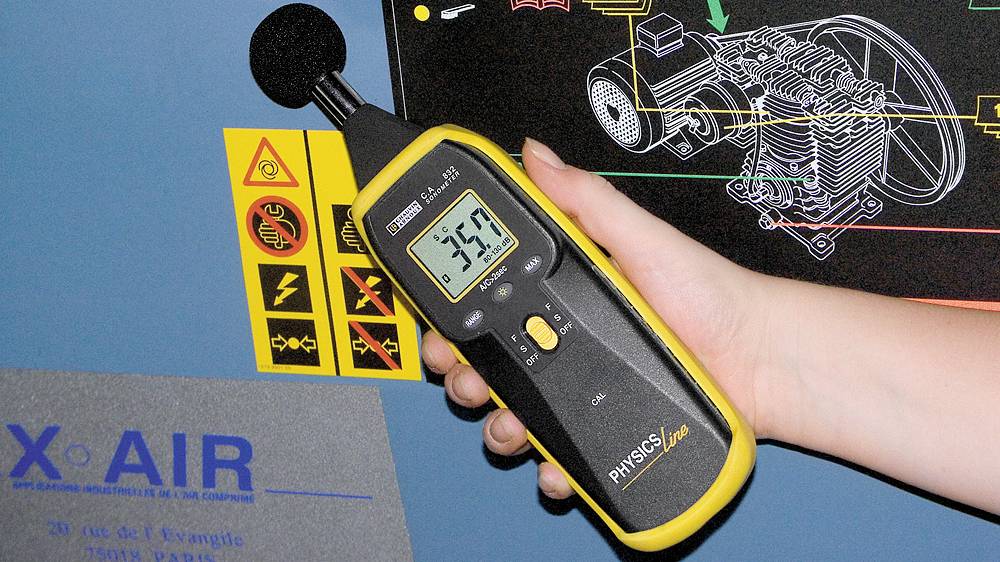 A hand is holding a sound level meter with a display showing '39.3'. Technical diagrams are visible in the background.