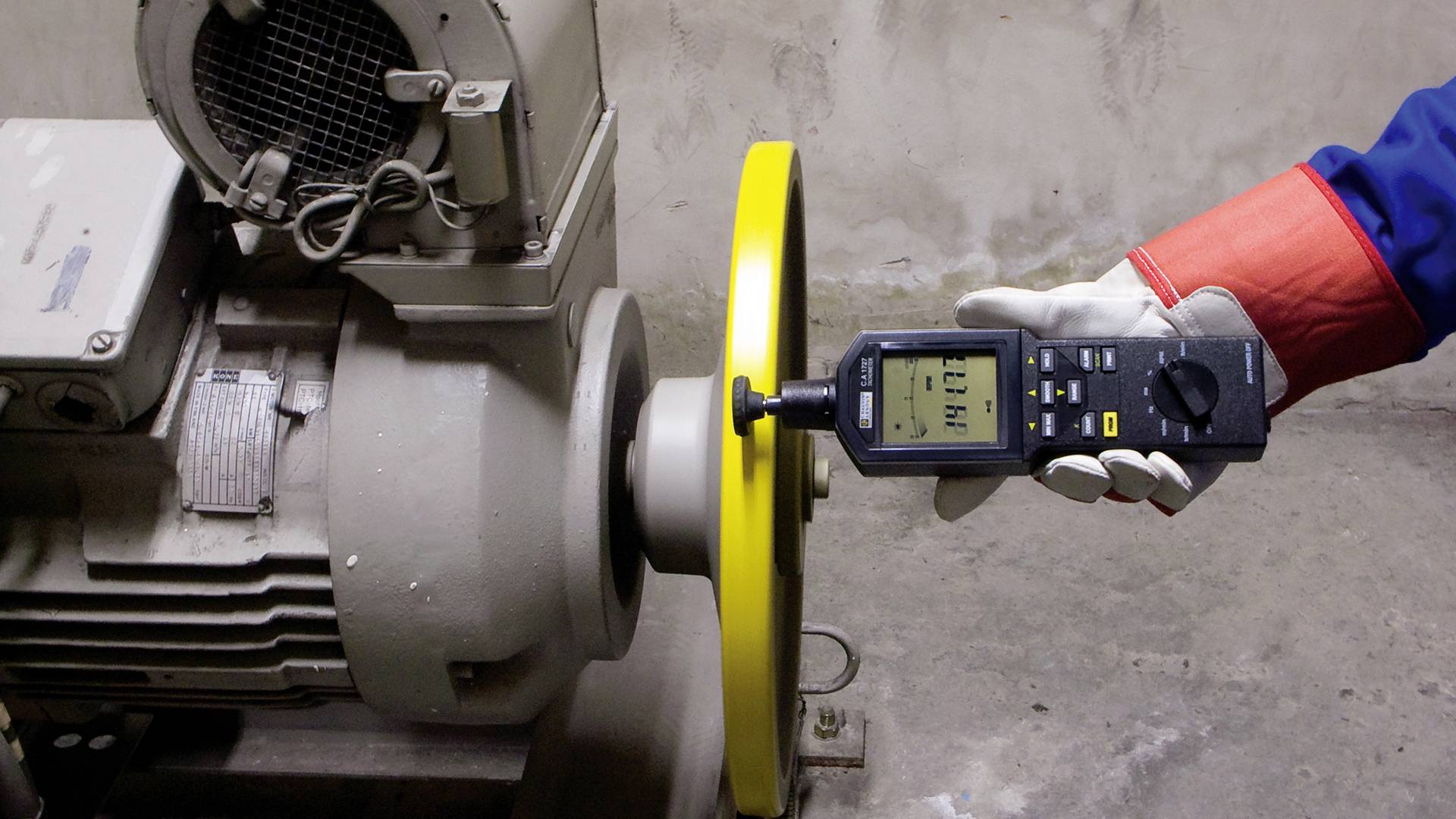 A person is measuring the vibrations of an electric motor using a vibration measuring device. The motor has a yellow protective guard.