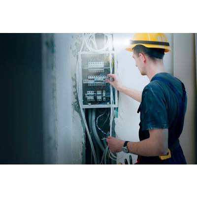 An electrician wearing a hard hat is working on an open electrical cabinet, checking cable connections, in an unfinished wall section.