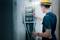 An electrician wearing a hard hat is working on an open electrical cabinet, checking cable connections, in an unfinished wall section.