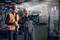 Two workers in hard hats and high-visibility vests are standing in a factory hall, one pointing at a machine, the other holding a document.