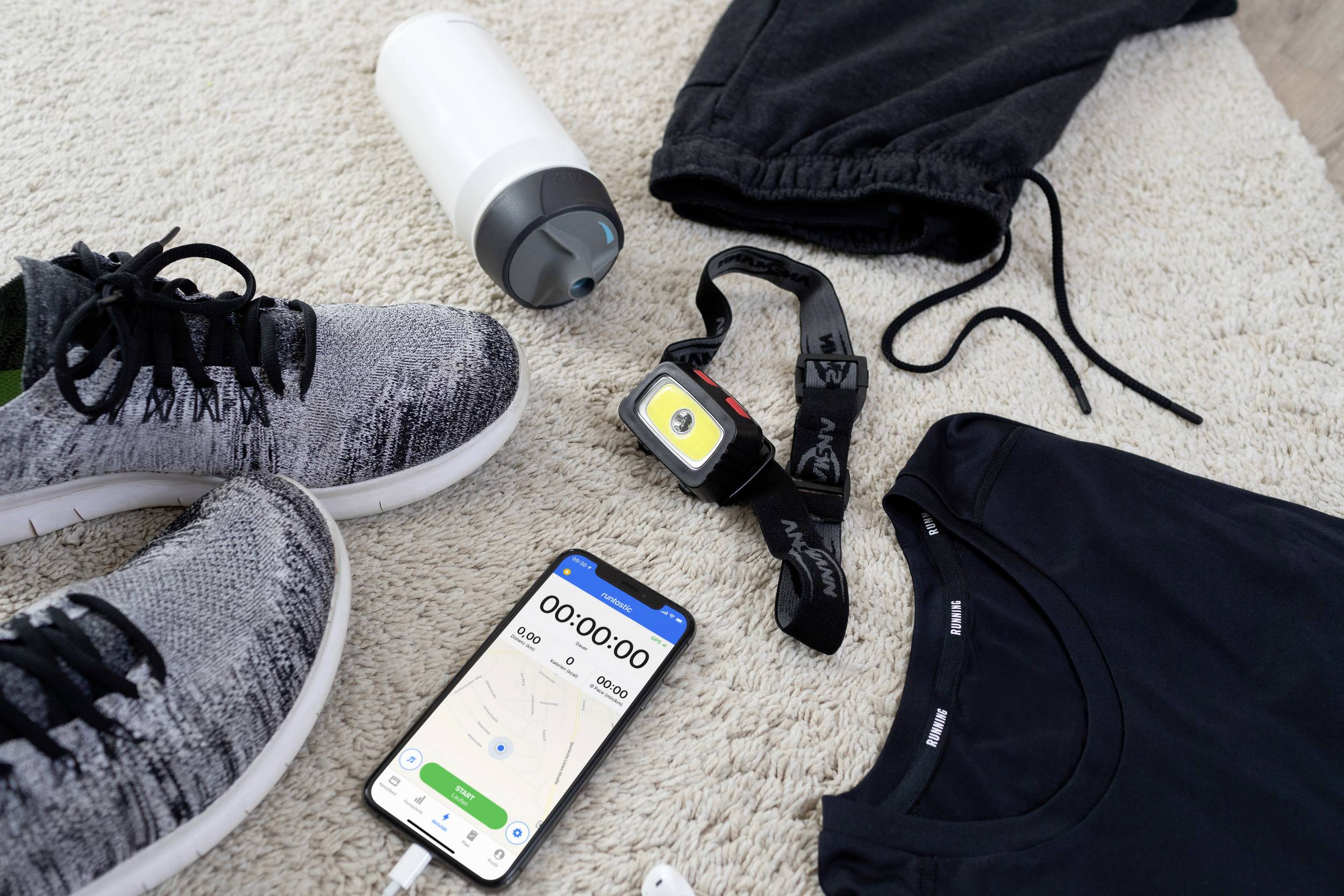 Sports equipment is laid out on a carpet: running shoes, sportswear, water bottle, fitness watch and a smartphone with a stopwatch app.