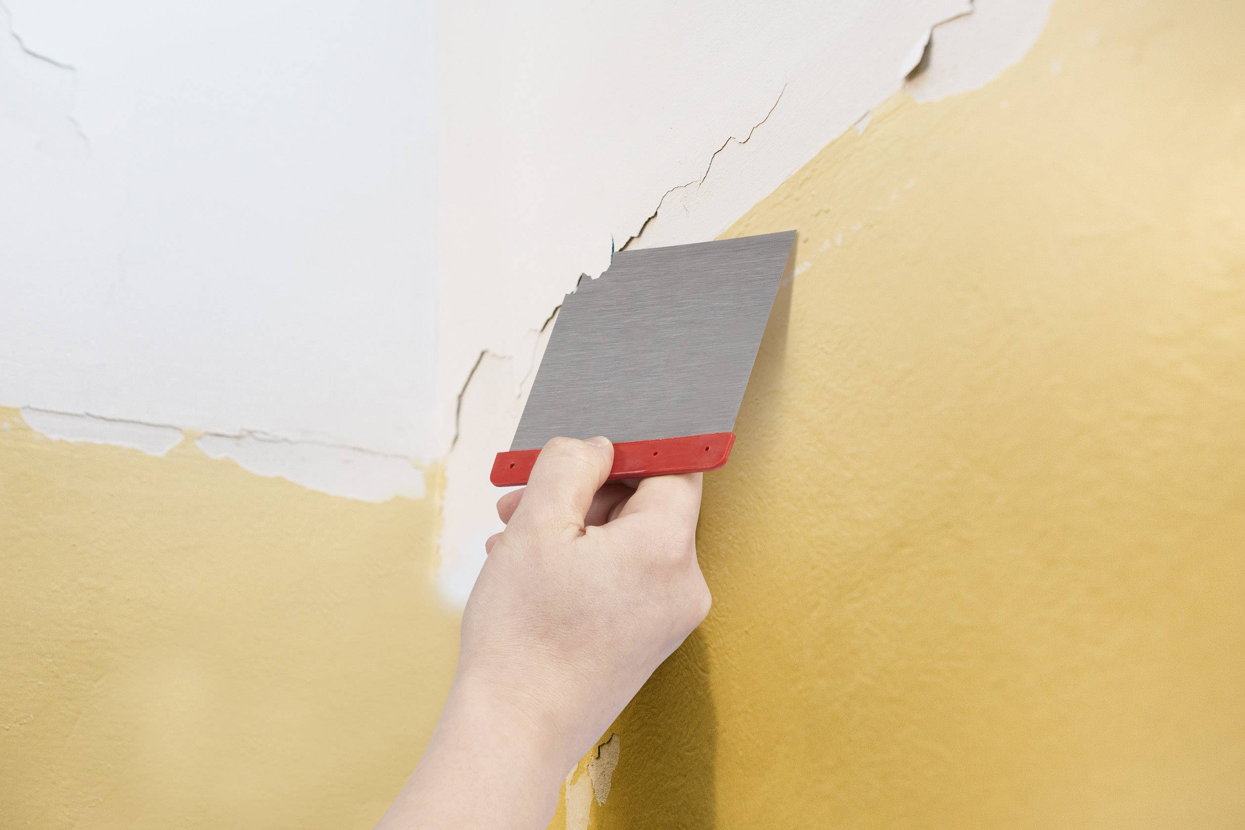 A hand with a plastering trowel is repairing cracks in a yellow-painted wall in the corner of a room.