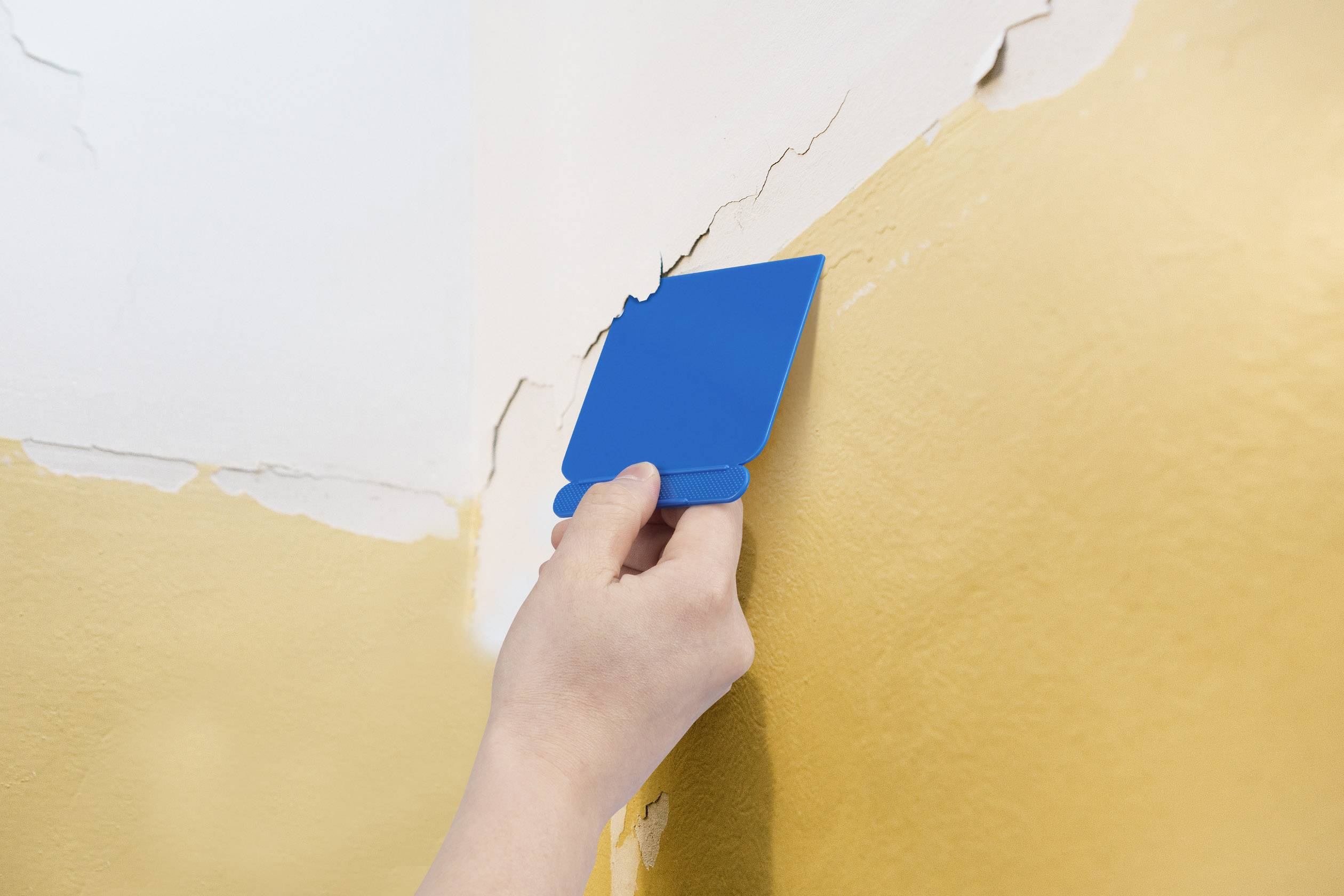 A hand holds a blue scraper, removing cracks from a wall painted yellow.