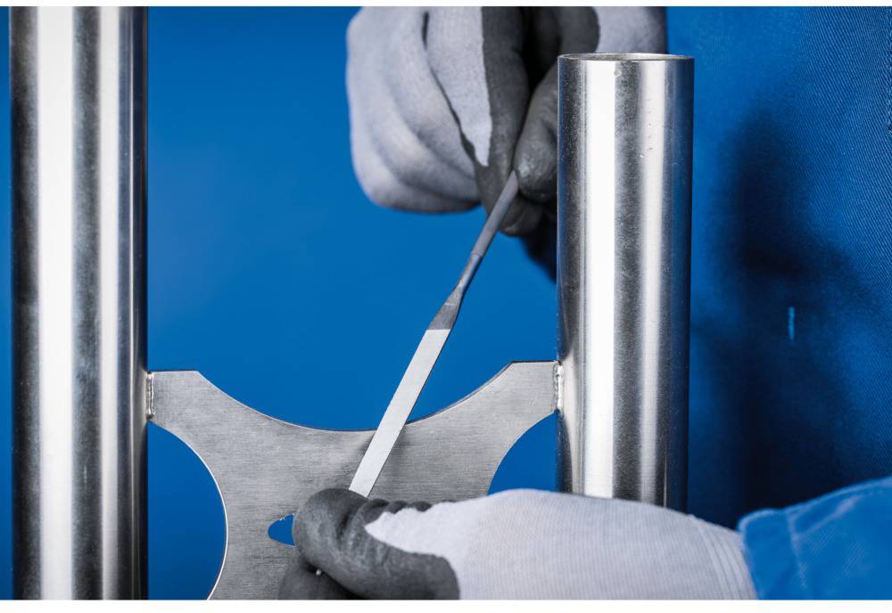 A man wearing gloves is filing a piece of metal, surrounded by two metal pipes.