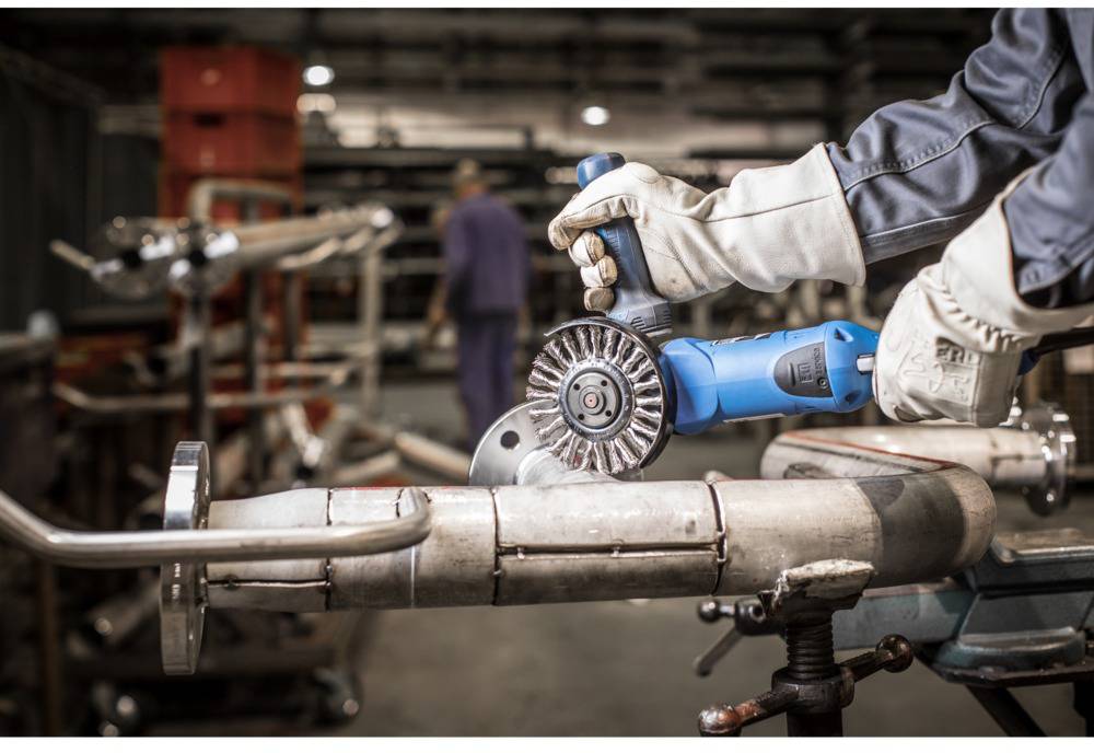 A person wearing protective clothing is grinding a metal pipe with a blue angle grinder in a workshop.