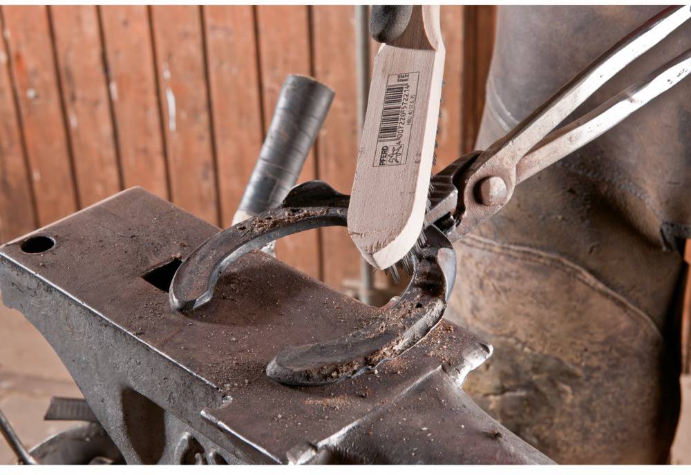 A farrier is shaping a horseshoe on an anvil. A pair of tongs holds the horseshoe, while a hoof knife trims the edges.