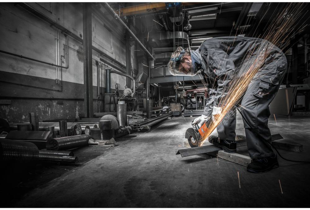 A person in protective clothing is grinding metal in a workshop, with sparks flying. Industrial machinery is visible in the background.