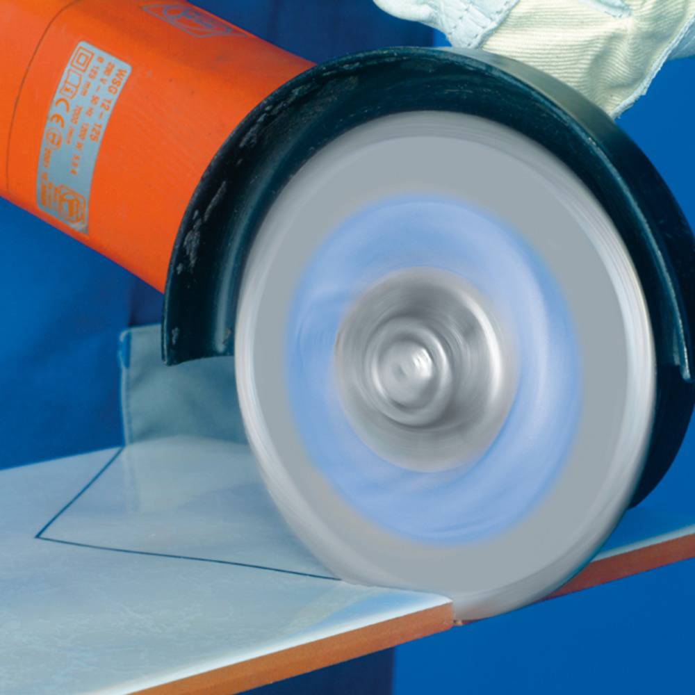 A red-orange electric grinding machine is grinding metal. A worker is wearing blue clothing and green protective gloves.