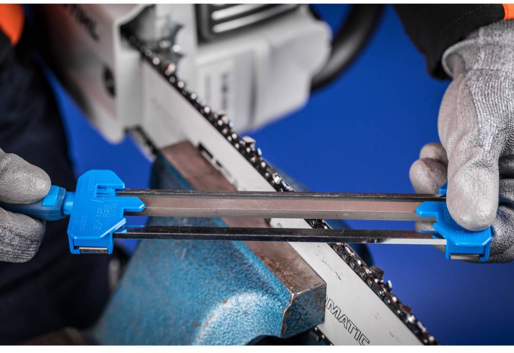 A person is sharpening a chainsaw with a file holder. The chain is clamped in place while the person wears work gloves.