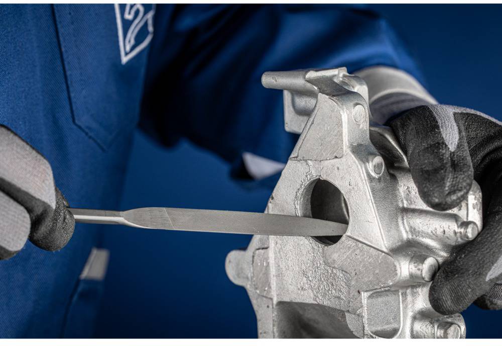 A person in workwear is using a file to work on a metal component. The focus is on precision and manual craftsmanship.