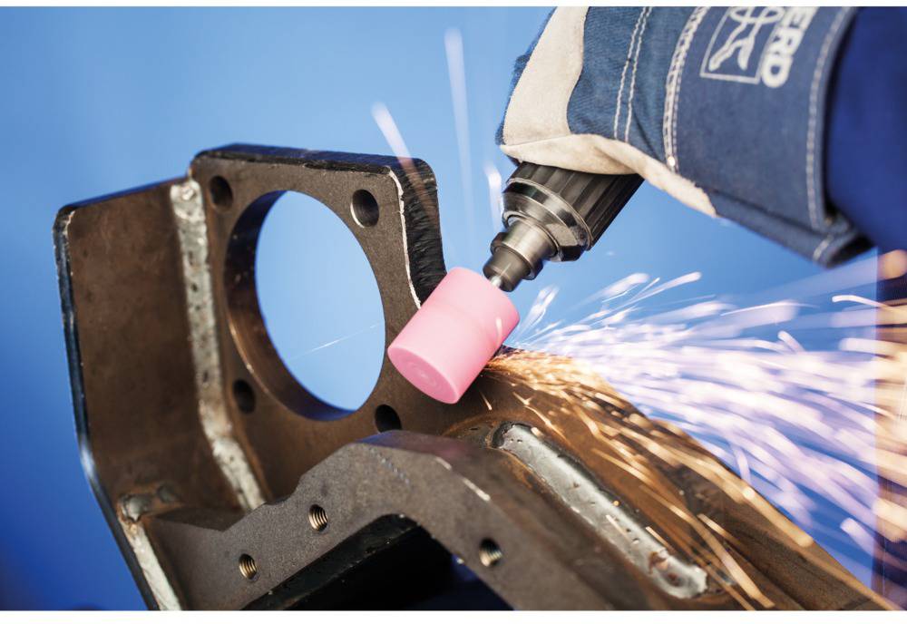 A hand wearing a work glove grinds a metal part with an electric grinding machine, sparks flying.
