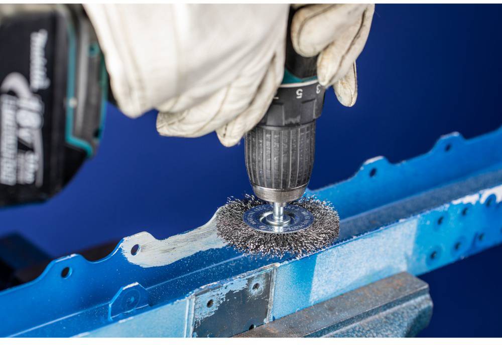 A person is using a power drill with a wire brush attachment to remove paint from a blue metal piece.
