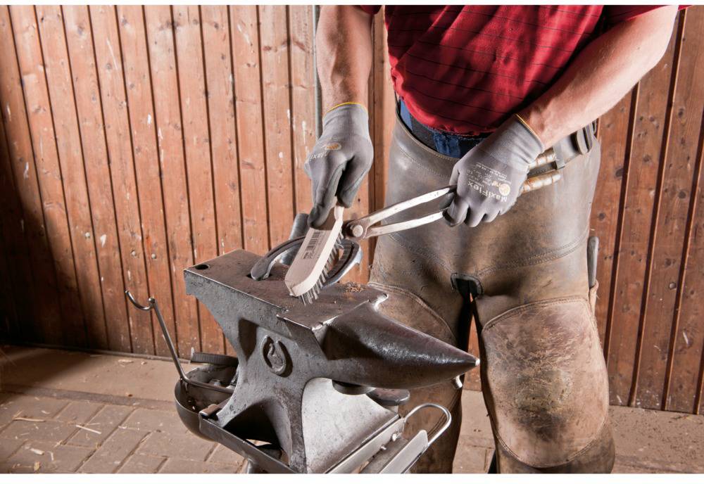 A blacksmith is shaping a horseshoe on an anvil in a workshop. He is wearing protective clothing and holding the horseshoe with a pair of tongs.