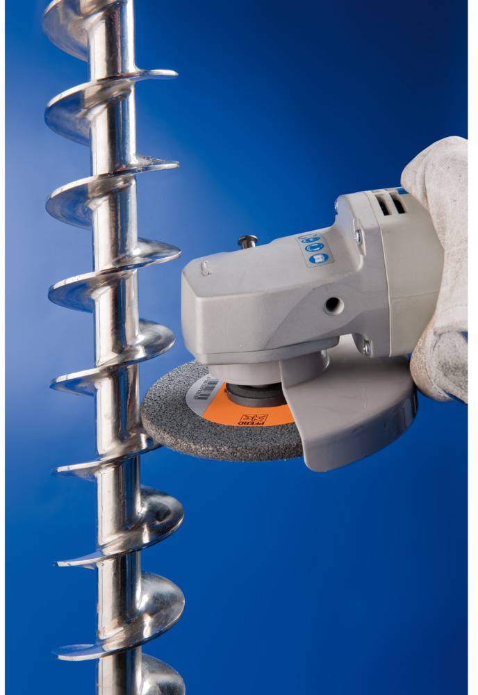 A hand holds an angle grinder working on a metal spiral against a blue background. Focus on the grinding tool and spiral structure.