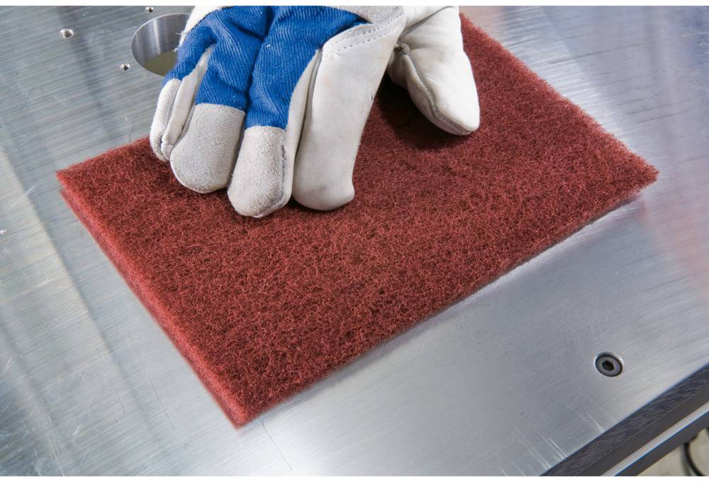 A hand covered with work gloves holds a red polishing pad on a smooth metal surface.