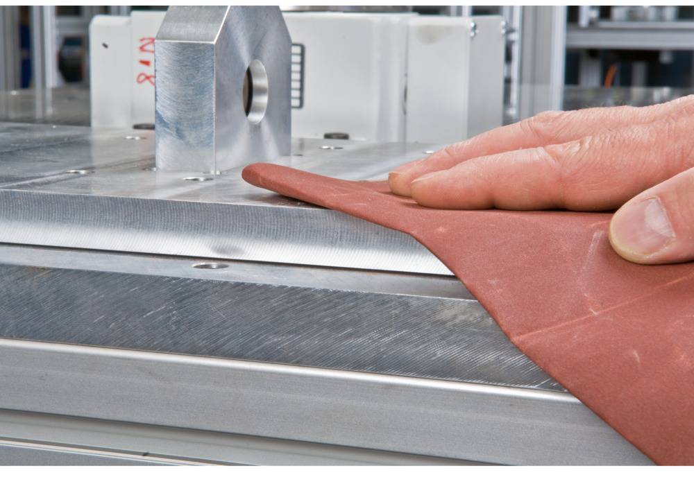 A hand holds a red piece of sandpaper and sands the edge of a metal plate. The background shows an industrial environment.