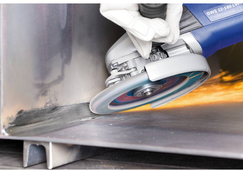 A person is grinding metal with an angle grinder, sparks flying. The grinder is blue, and the person is wearing white protective gloves.