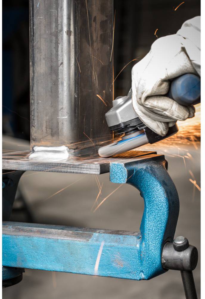 A person wearing a glove is grinding a metal edge with an angle grinder, sparks flying. Metal clamped to a workbench. Industrial environment.
