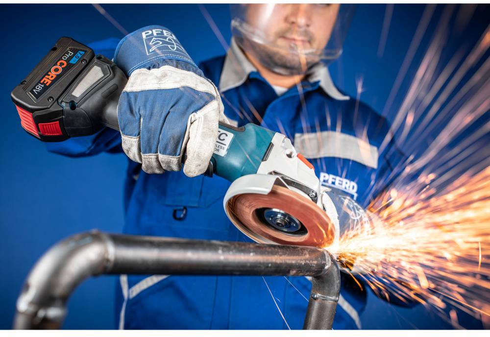 A worker in a blue uniform is grinding a pipe with an electric grinding machine. Sparks are flying from the grinding wheel.