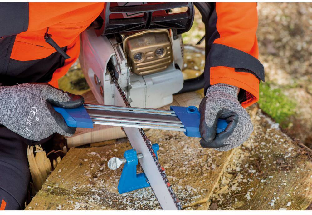 A person is sharpening the chain of a chainsaw with a chainsaw sharpening kit. The person is wearing an orange protective jacket and grey gloves.