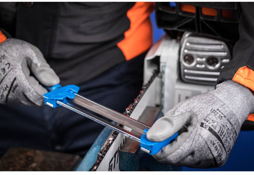 A person wearing gloves is sharpening the chain of a chainsaw with a blue file.