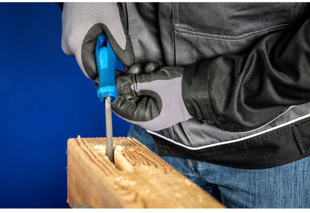 A person in workwear is filing a rectangular hole in a wooden board. They are wearing gloves and using a metal file.
