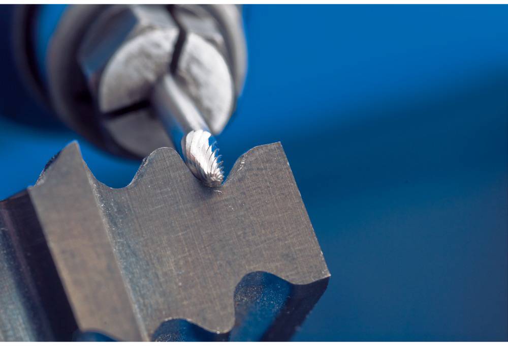 A drill shapes a serrated metal piece. The scene depicts the precision process of metalworking in a workshop.