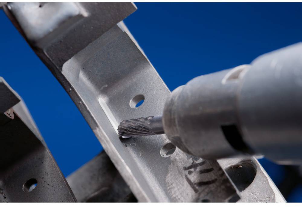 Close-up of a metal drill bit drilling through a metal piece with multiple holes. The blue background emphasises the action.