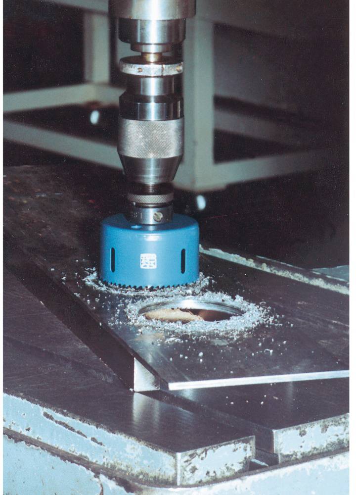 A blue hole saw is attached to a drill and is drilling a round hole into a metal plate on a workbench.