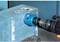 A blue hole saw cuts a round hole in a grey metal enclosure. A black drill is attached to the saw.