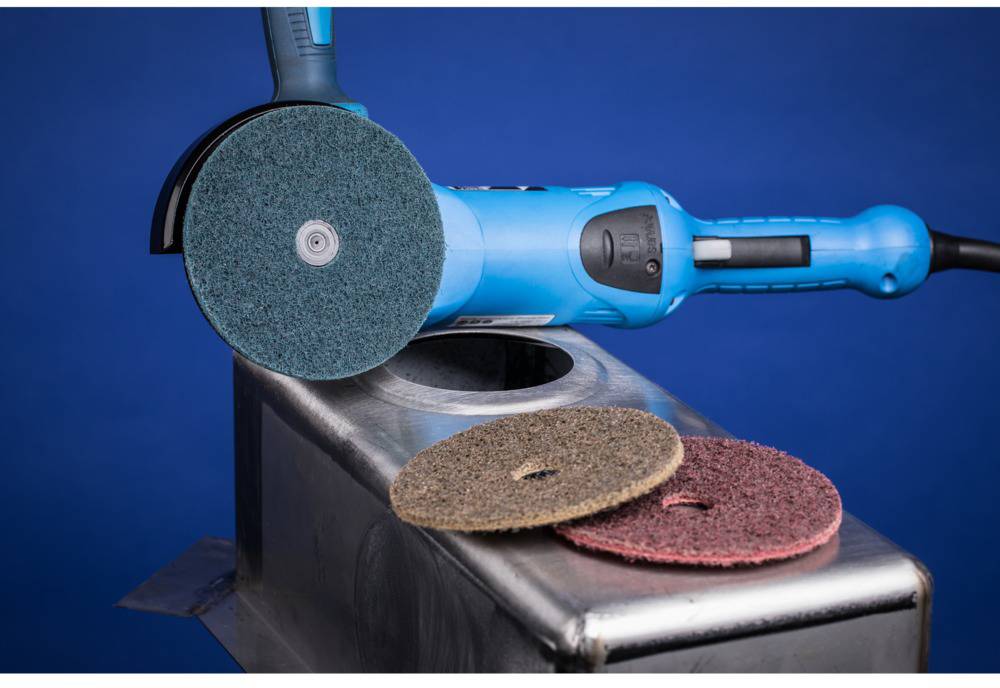 An angle grinder with an attached grinding pad is lying on a metal container. Two additional grinding pads are placed beside it.