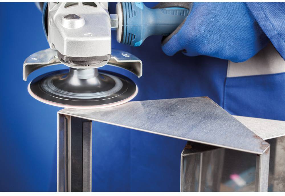 A craftsperson grinds a metal plate with an angle grinder, sparks flying. The person is wearing blue protective clothing, with a blue background.