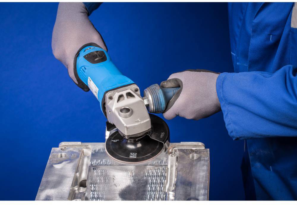 A person in blue workwear is grinding a metal surface with an angle grinder. Hands are wearing safety gloves.