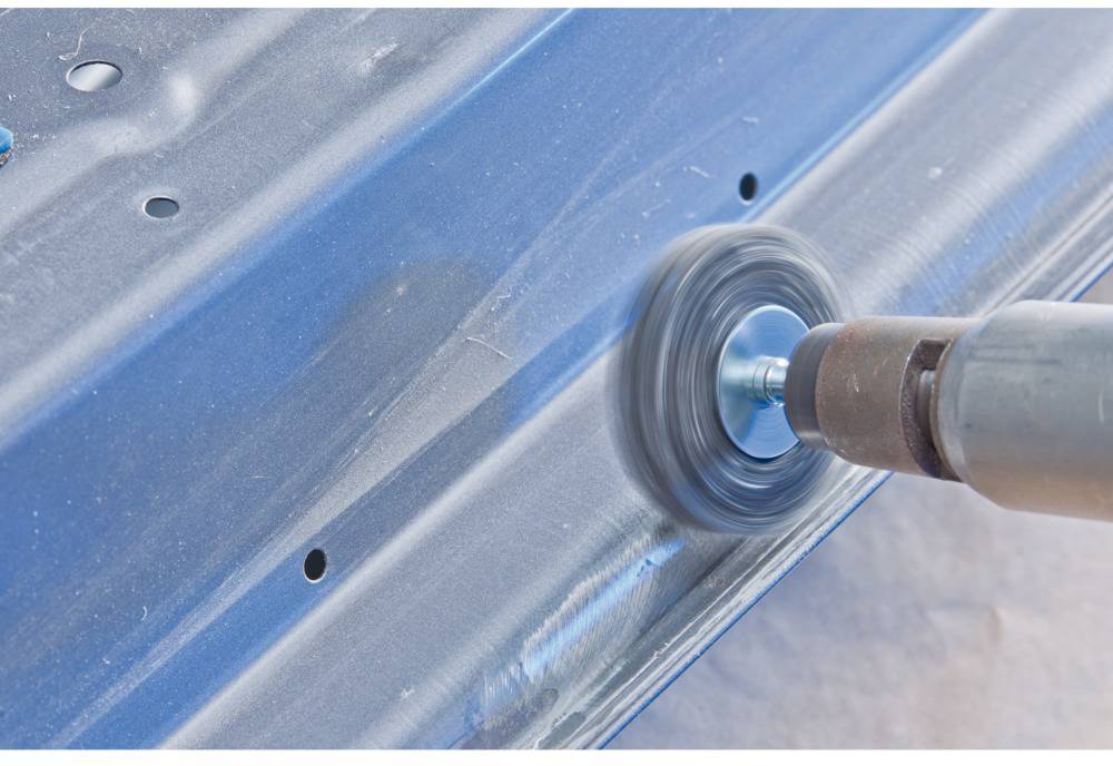 A rotating wire brush cleans the surface of a blue metal sheet, removing rust or dirt for industrial applications.