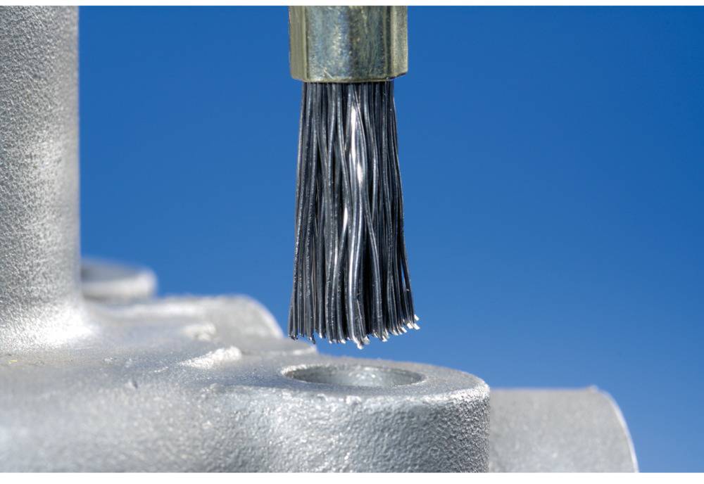Close-up of a wire brush head in front of a blue-grey metallic object, likely used for cleaning or processing surfaces.