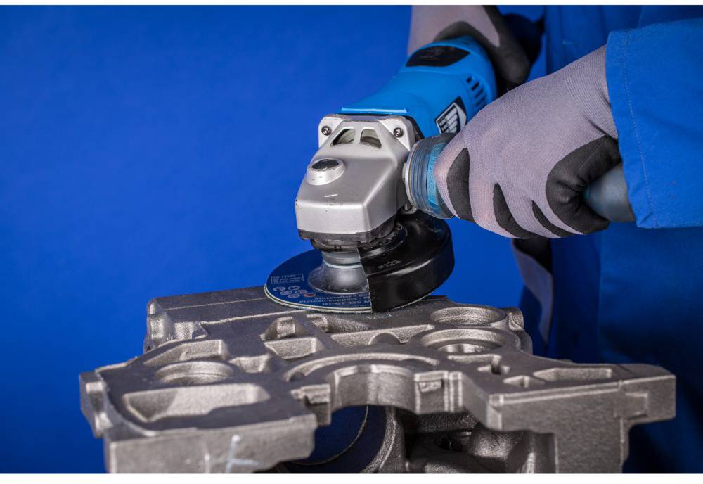 A person in workwear grinding a metal piece with a blue angle grinder against a blue background.