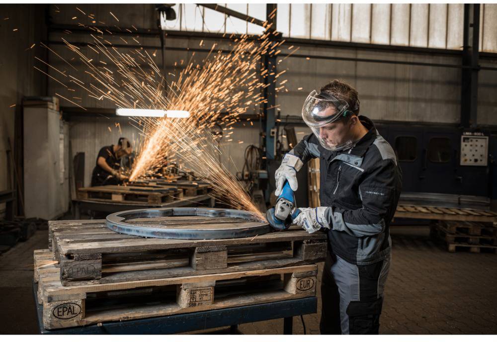A worker wearing safety glasses is grinding metal in a workshop, with sparks flying. Another worker is visible in the background.