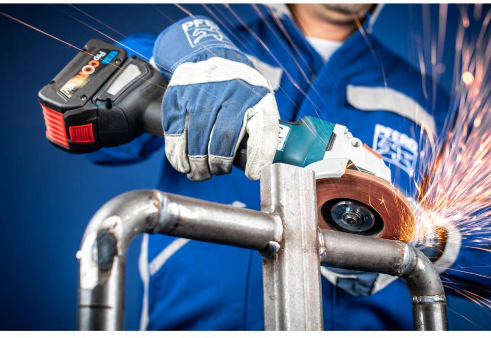 A person in blue workwear is grinding a metal pipe with an angle grinder, with sparks flying.
