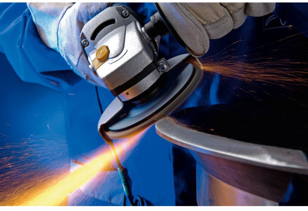 A worker is grinding metal with an angle grinder, sparks flying. He is wearing blue protective clothing and gloves.