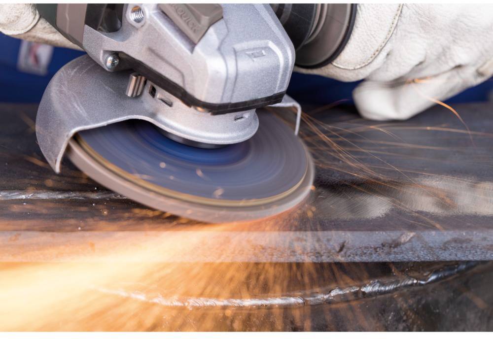 A close-up of an angle grinder spraying sparks as it glides over a metal surface. Worker is wearing protective gloves.