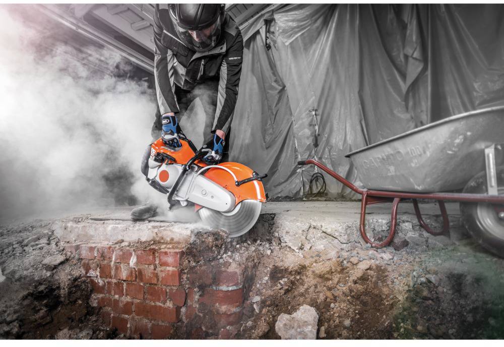 A worker in protective equipment is using an electric chainsaw to cut through a brick wall on a construction site.