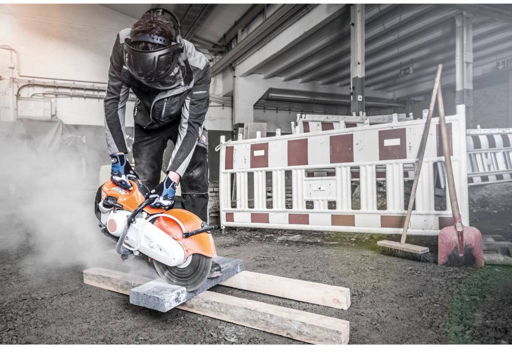 A person in protective clothing is cutting metal with a chainsaw in an industrial environment. Barriers are visible in the background.