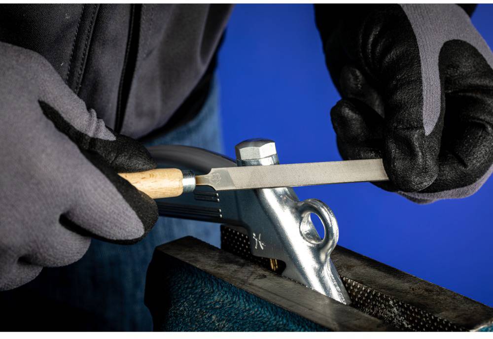 A person is filing a spanner in a vice with a metal file tool, wearing black protective gloves.