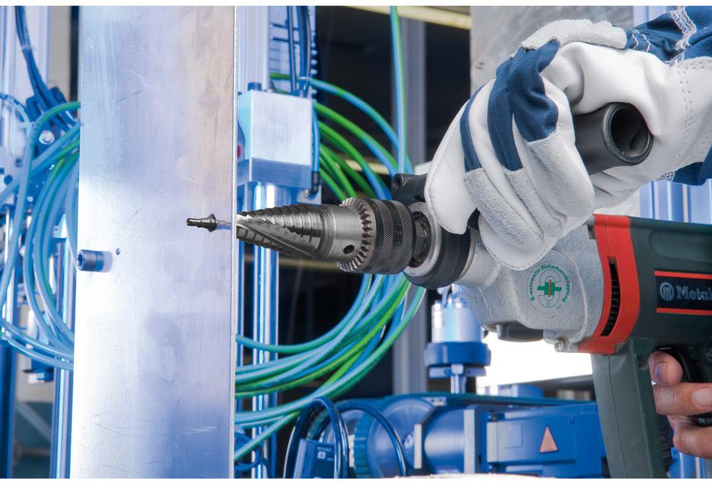 A person is drilling into a metal piece in an industrial setting. Green cables and blue machinery in the background.