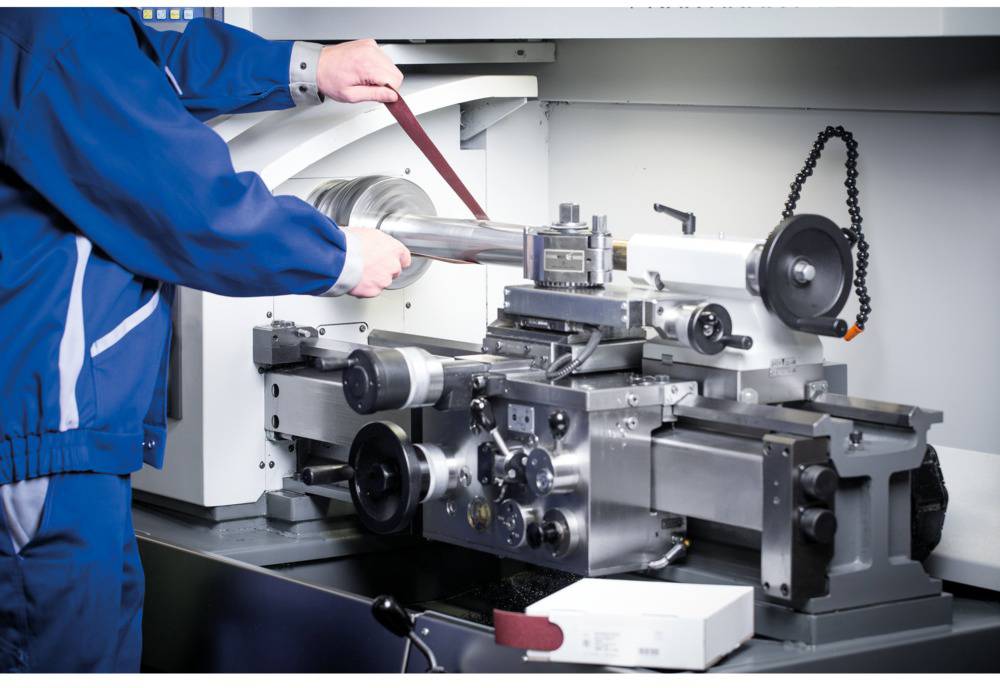 A person is machining a metal workpiece on a lathe in a factory, wearing blue overalls and using a grinding belt.