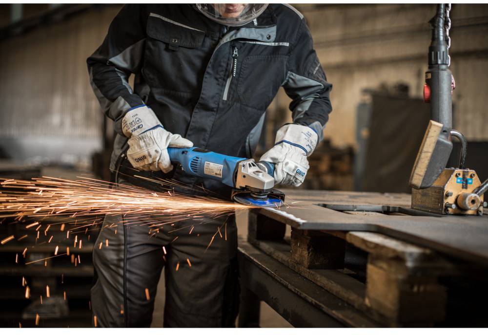 A person in protective clothing is cutting metal with an angle grinder, with sparks flying. They are in a workshop environment.