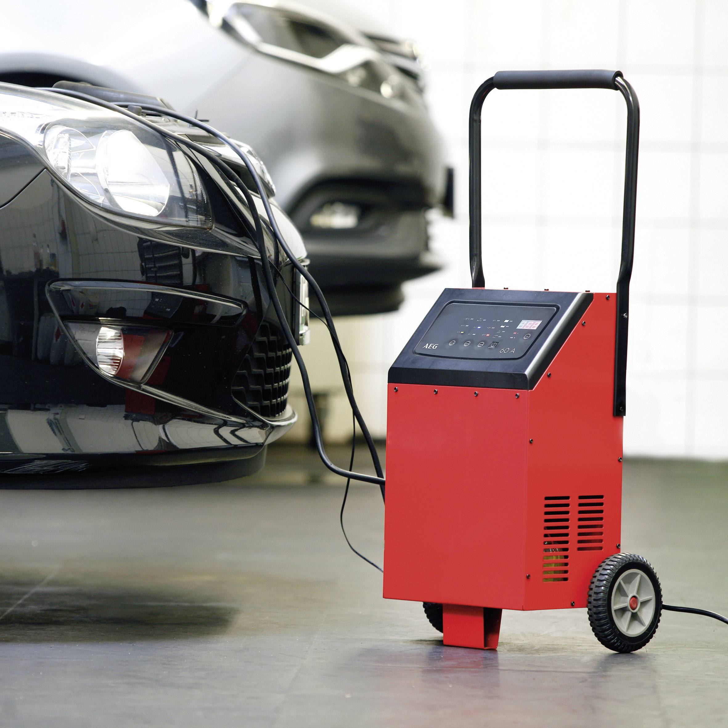 A black car is being charged in a garage with a red battery charger. The charger is standing on two wheels.