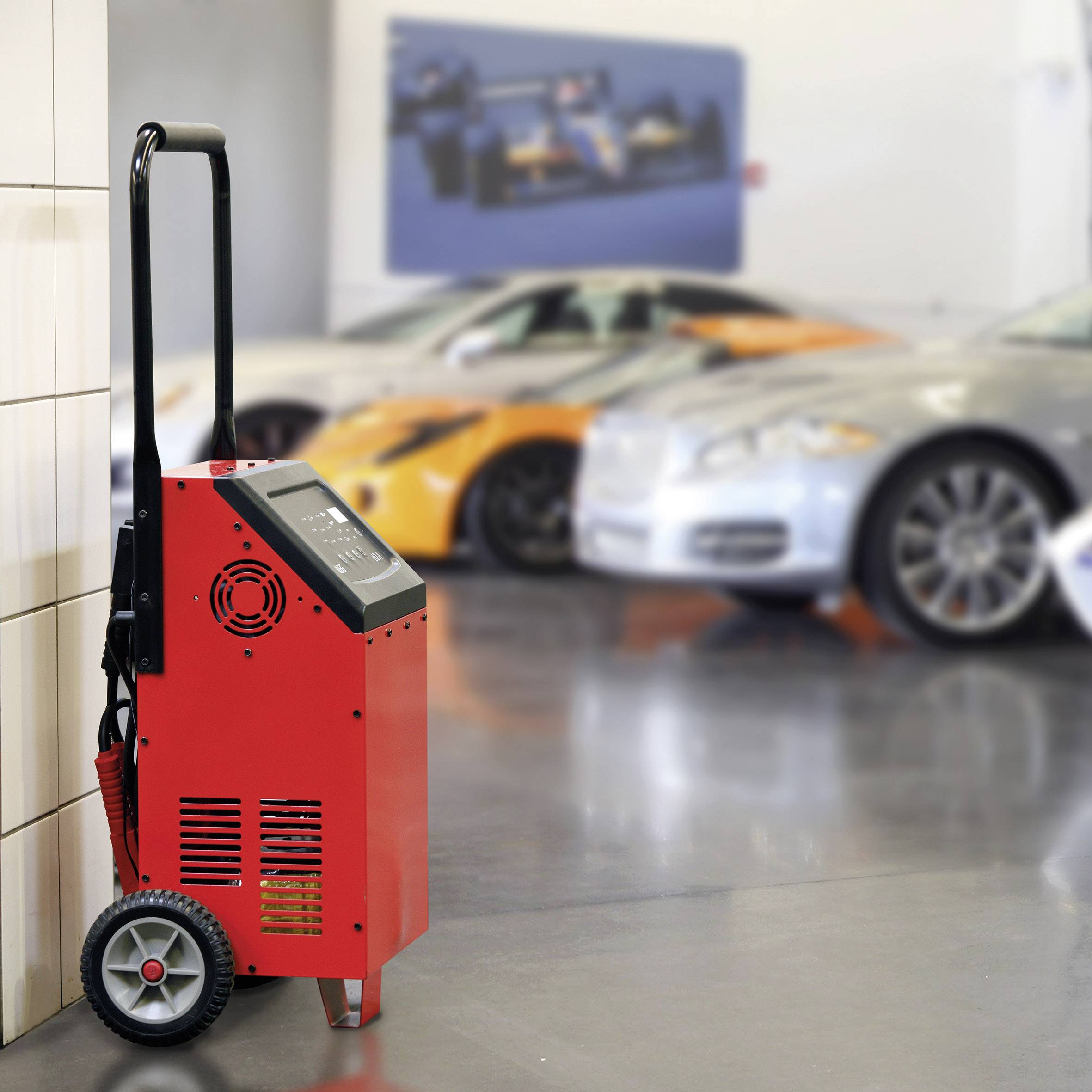 A red battery charger on wheels stands in front of blurred sports cars in a garage.